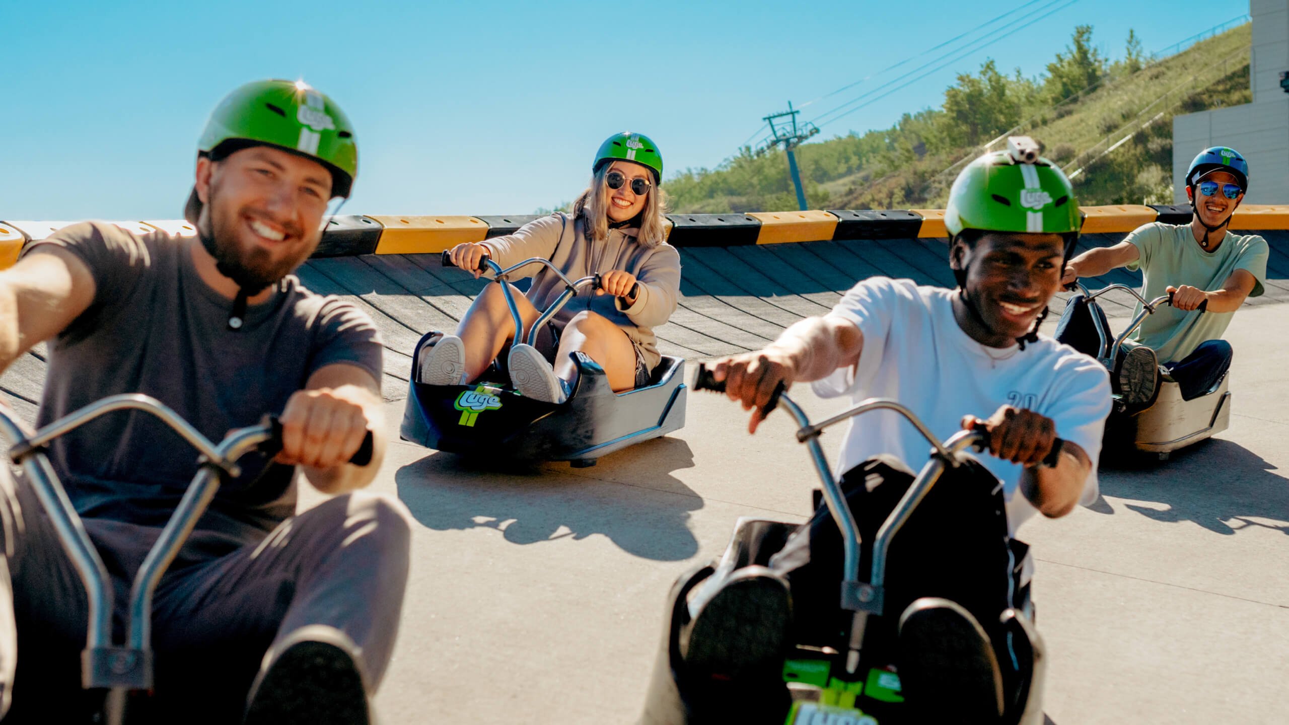 Four friends smiling in the sun as they turn a wide corner on a luge track